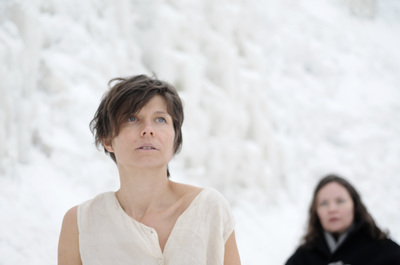 Nainen seisoo lumisen ja jäisen kallion edessä ohuessa paidassa, takana seisoo toinen nainen.