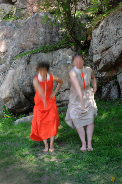 Kaksi naista seisoo kallion edessä nurmikolla vasen käsi lanteilla ja oikea suorana eteenpäin. 