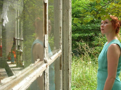 Nainen seisoo kesäisessä pihassa vanhan puutalon ruutuikkunan edessä.